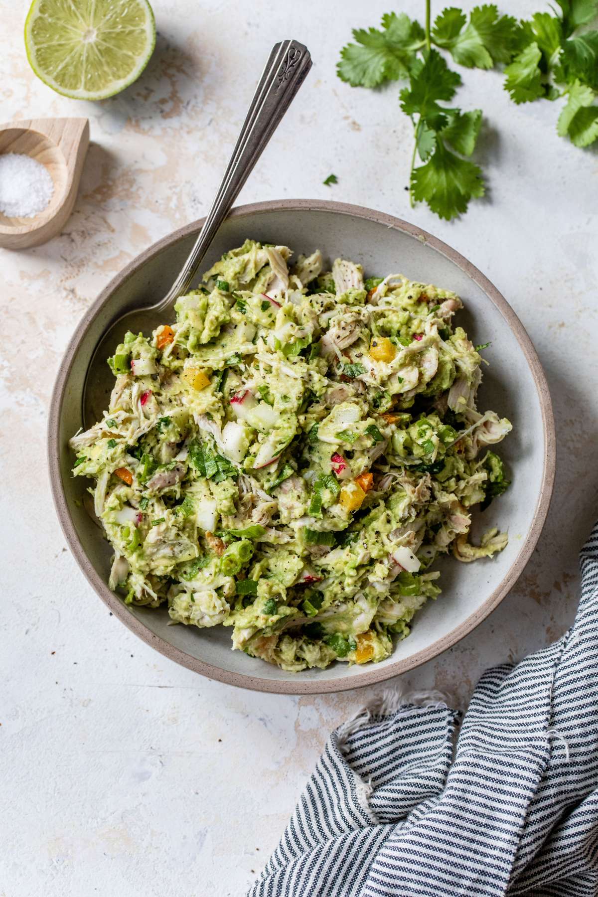 Chicken salad mixed with avocado and veggies in a bowl near fresh cilantro and lime.