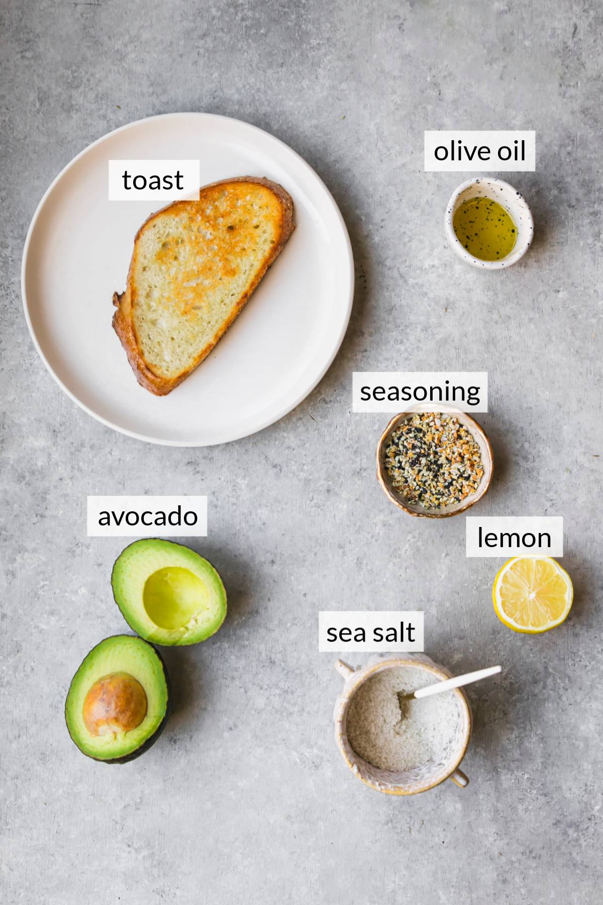 Ripe avocado, a piece of toast on a plate, half of a lemon and a bowl of seasoning.