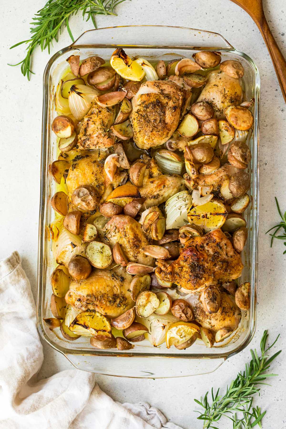 Chicken and potatoes in a glass baking dish near fresh rosemary.