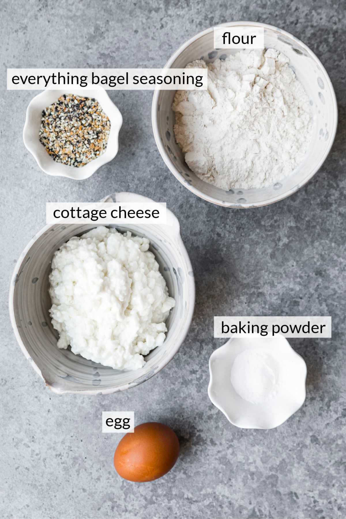 An egg near bowls of cottage cheese, flour, baking powder and seasoning.