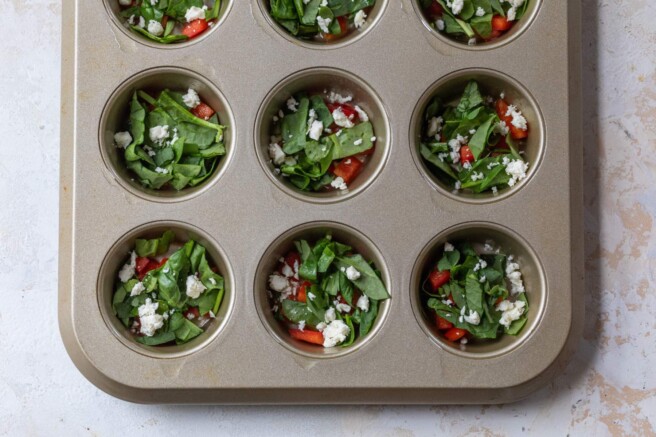 Adding spinach, diced red bell pepper and feta cheese to a muffin pan.