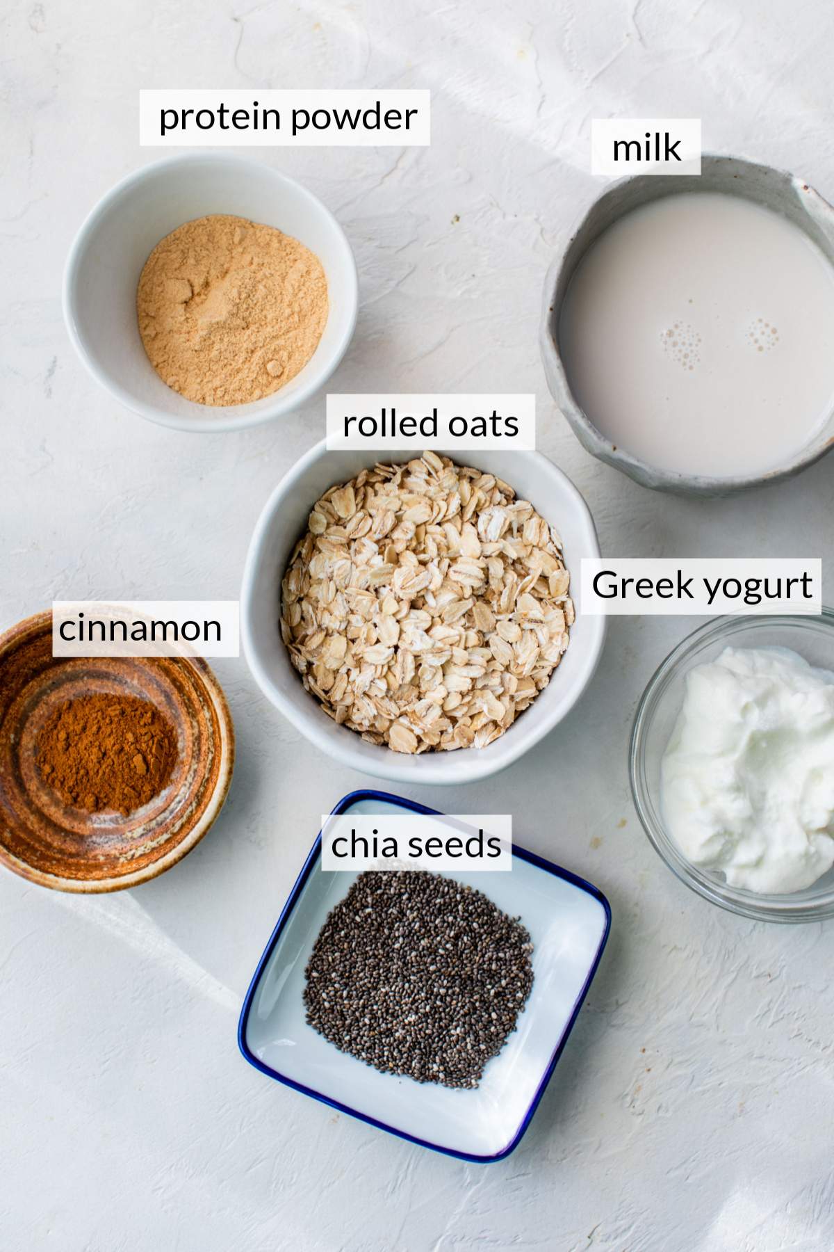 Small bowls with rolled oats, milk, protein powder, chia seeds and greek yogurt.