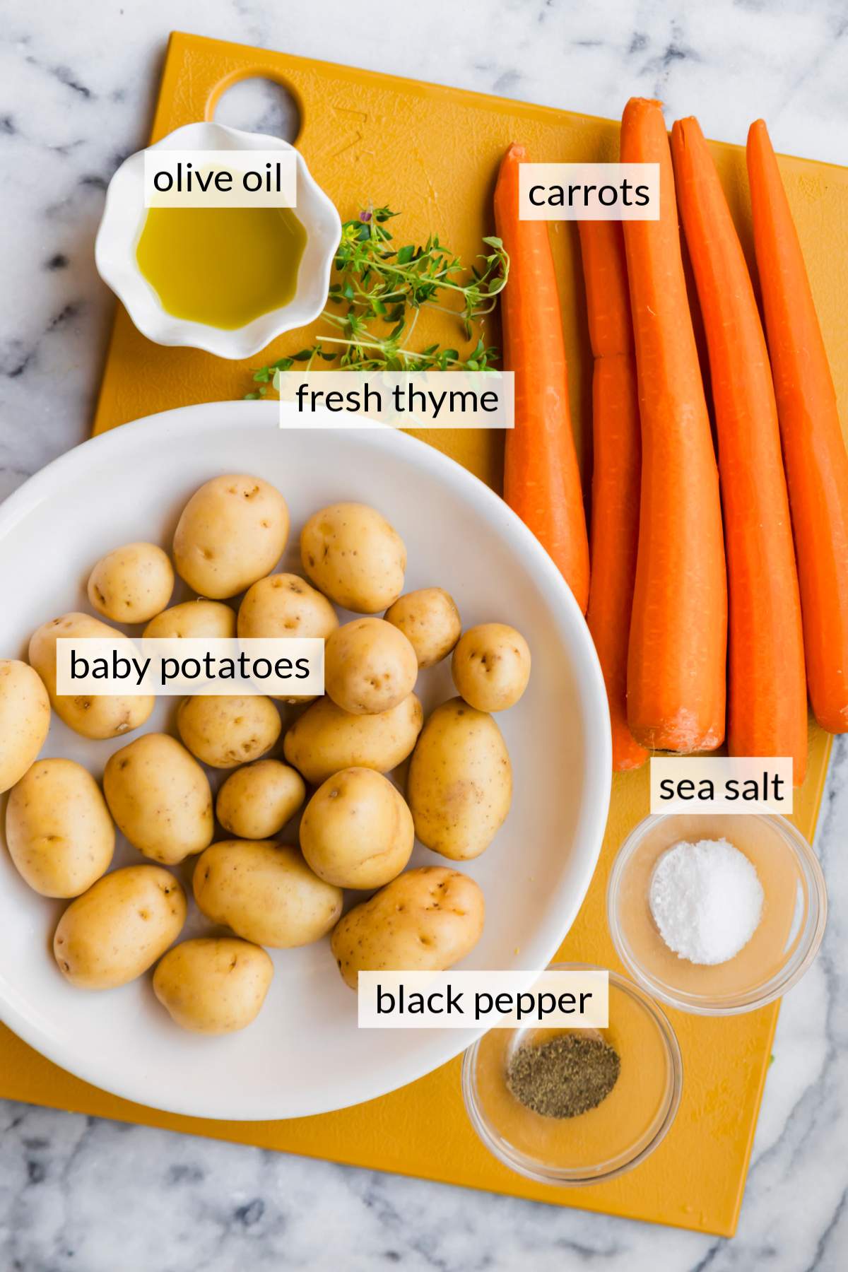 Bowl of potatoes near carrots, thyme, salt and pepper on a cutting board.