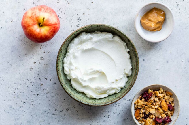 Greek yogurt in a bowl near an apple, a small bowl of peanut butter and a bowl of granola.